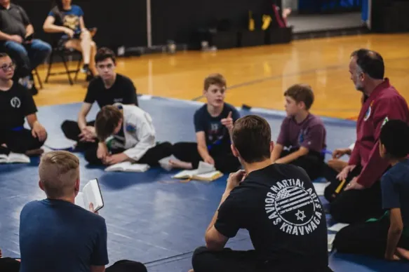 Students seated in a circle on the mats with their Bibles open for a devotional, an instructor in a red gi among them